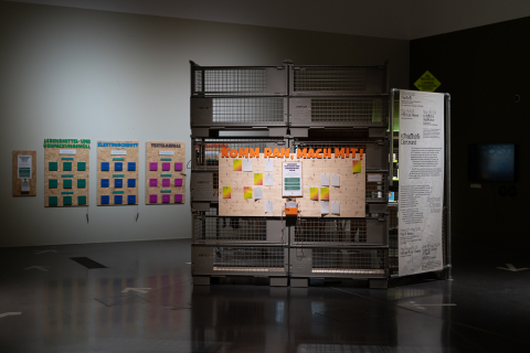 Kunstinstallation in einer Ausstellungshalle zum Thema Kreislaufwirtschaft und Repairkultur: Metallgitterboxen mit Holzpaneelen zeigen Schilder mit Aufschriften wie "Lebensmittel- und Verpackungsmüll", "Elektroschrott", "Textilabfall" und "Komm ran, mach mit!". Der Raum ist dunkel mit Spotlights, der glänzende Boden spiegelt die Exponate.